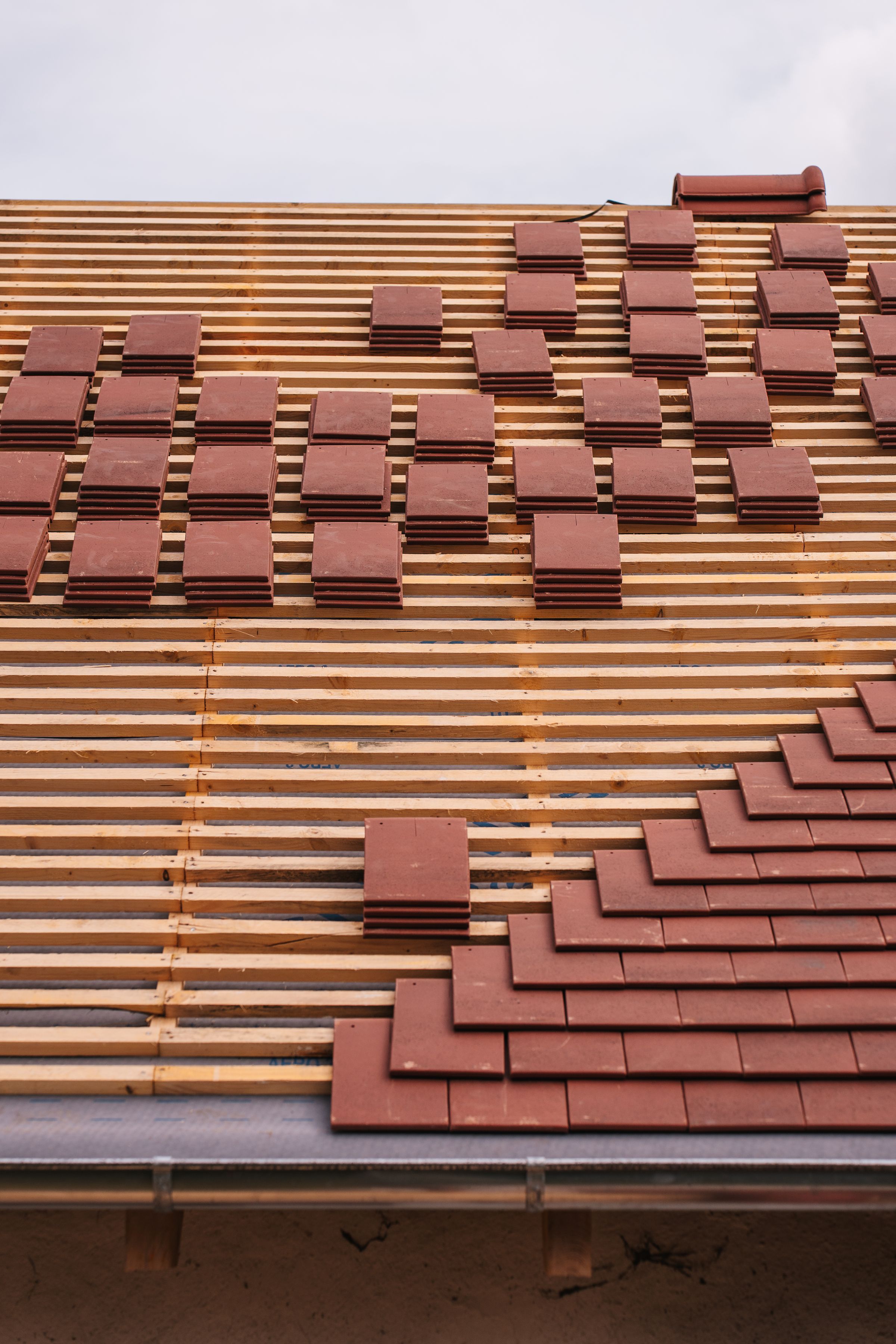 Rampant de toiture en cours de couverture : liteaunage cloué sur écran de sous-toiture et piles de tuiles plates réparties avant la pose